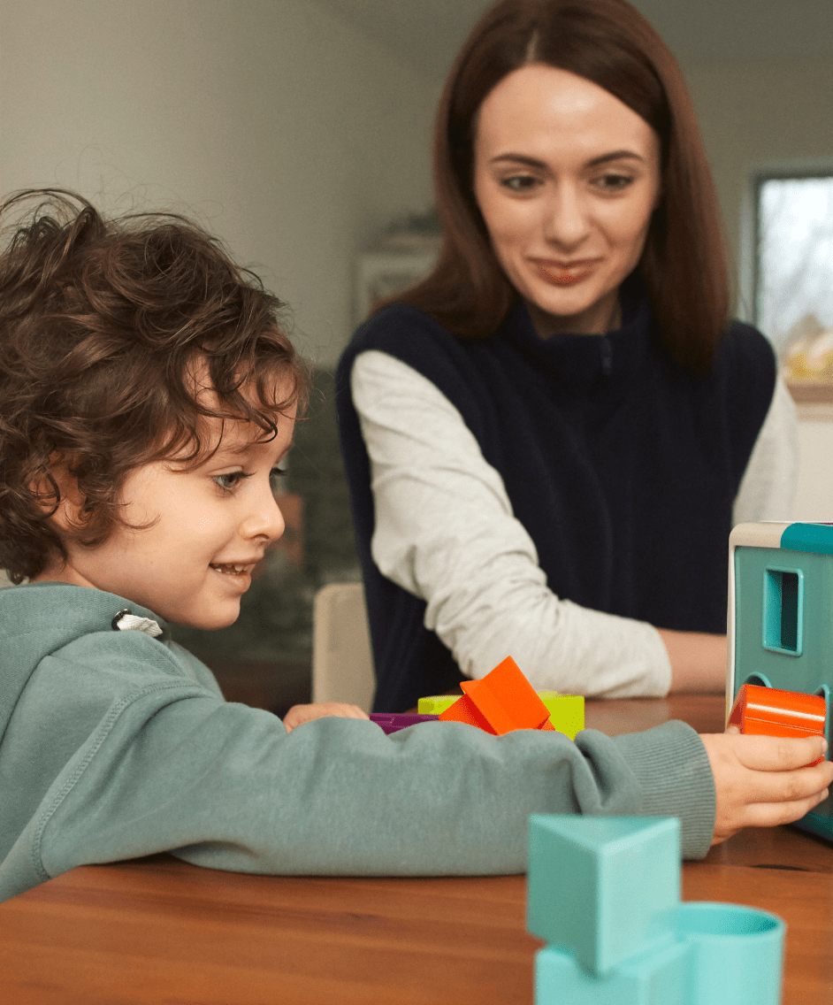 A white child with brown hair playing with blocks, while a white woman observes the behavior